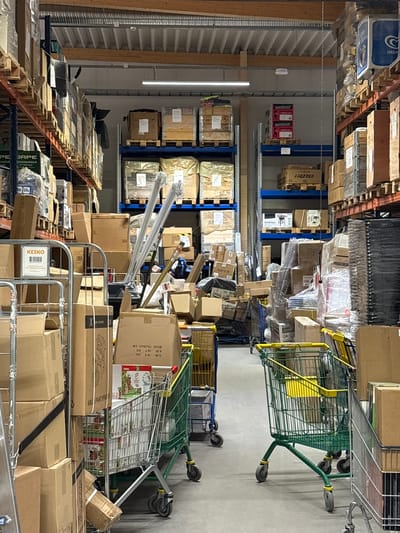 This media file depicts an indoor view of a warehouse or retail storage area filled with numerous boxes and shelving units. The scene is dominated by cardboard boxes of various sizes, many stacked on wooden pallets and metal shelving. Some boxes are neatly arranged, while others appear to be in disarray, suggesting active storage or movement of goods.
Visible objects include several shopping carts, one of which is green and yellow, and another is made of metal with wheels. One of the shopping carts contains what appears to be a stack of printed materials or packaging. A prominent piece of text visible on a box reads "KESKO," a Finnish retail cooperative. Other visible text includes "Collector," "JAPE," "POHONKERÄÄJÄ," "FREZZER," and "INGM," which likely refer to product names or brands.
The setting appears to be a well-lit industrial or commercial space, with fluorescent lighting visible in the background. The overall impression is one of a busy, functional storage facility. No people are visible in the frame. The time of day and weather conditions are not discernible from the image, but the indoor setting suggests the activities are independent of external environmental factors. The location context of Pori, Finland, suggests this is likely a retail distribution center or a large stockroom for a Finnish business.