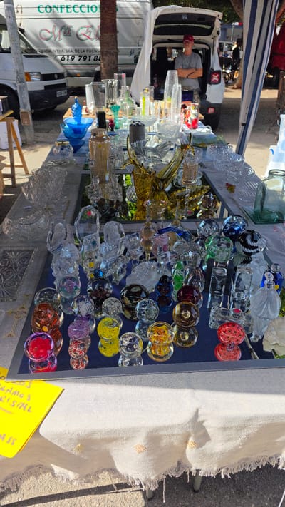 A high-angle medium shot shows a street vendor's table laden with a variety of glassware, displayed outdoors. The table is covered with a white cloth that has frayed edges and is adorned with embroidered yellow and orange accents. To the left, a bright yellow sign with handwritten text in Spanish reads "OFRECEMOS CRISTAL" ("We offer crystal"). Behind the table, a white van with Spanish text and a phone number "619 791 88" is visible, suggesting the vendor might be operating out of it. A man wearing a red baseball cap and a grey t-shirt stands with his arms crossed behind the open back of a white vehicle, possibly a Jeep, which is parked directly behind the table. The scene is set in Almuñécar, Spain, as indicated by the presence of Spanish text and the general appearance of a street market environment. The background reveals trees and other market stalls, suggesting a bustling outdoor market. The glassware on display includes a diverse assortment of clear and colored glass items, such as bowls, vases, figurines, and decorative spheres.