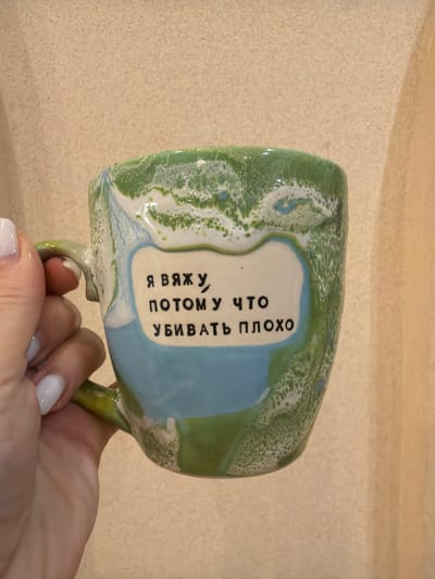 A hand holds a green and blue glazed ceramic mug with the Russian text "Я вяжу, потому что убивать плохо" ("I knit, because killing is bad"). The background appears to be a light beige wall. There is no information to definitively place this image in Levkovo, Russia.