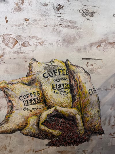 A mural on a textured, off-white wall depicts three burlap sacks overflowing with coffee beans. The sacks are rendered in shades of yellow, beige, and brown with dark outlines and streaks of orange and green. The sack in the center has black lettering that reads "COFFEE BEANS" and "Organico" underneath, along with what appears to be a date: "26-11-2019". Another sack on the left has "COFFEE BEANS Organico" written on it. A third sack, partially visible on the right, has the partial word "COF" visible. The bottom of the mural shows a pile of dark, reddish-brown coffee beans spilling from the sacks. The wall itself has a distressed, rough texture with patches of exposed brown material. This artwork is located in Epsom and Ewell, United Kingdom.