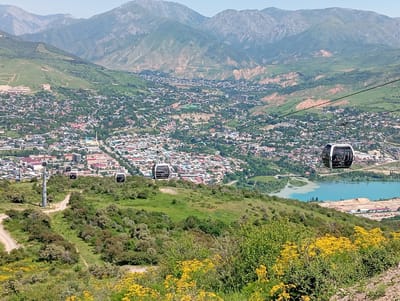 The image captures a panoramic view of the city of Chinarkent, Uzbekistan, set against a backdrop of rolling hills and distant mountains under a clear, bright sky. The foreground is dominated by lush green vegetation interspersed with patches of yellow flowers. A cable car system traverses the scene with several cars suspended mid-air. Below, the city unfolds with a mix of residential buildings and green spaces stretching across the valley. A turquoise-colored lake is situated on the right side of the scene. The cable cars, along with the lake, serve as defining elements, contrasting the natural landscape with human development.
