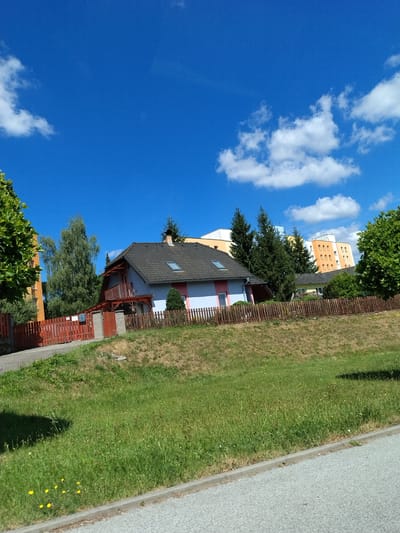 The image depicts a residential area under a clear blue sky with scattered white clouds. In the foreground, a paved road runs along the bottom edge, bordered by a strip of green grass with some small yellow wildflowers. Behind the grass, a sloped embankment is covered in a mix of green grass and dry, brown vegetation.
Dominating the middle ground is a light blue house with a dark grey tiled roof, featuring a wooden balcony with a railing, supported by wooden posts. A small dormer window is visible on the roof. The house is surrounded by a dark brown wooden fence. To the left of the house, a red wooden fence lines a driveway. Adjacent to the driveway, a brick post holds a mailbox.
Behind the house and fence, several tall green trees are visible, including a prominent birch tree with light-colored bark. In the background, two tall apartment buildings with orange and white facades stand out. The scene is consistent with a suburban or semi-urban environment, potentially in Volyně, Czechia, given the architectural style of the buildings and the overall landscape. No people or specific actions are evident in the image.