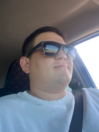 A medium shot captures a person wearing sunglasses and a white t-shirt while seated in a vehicle. The person is looking slightly off-camera to the right. The sunglasses reflect a road scene with a truck and a clear sky. The interior of the car is visible, including a black seat cover with red accents and a seatbelt. The overall lighting suggests it is daytime. The image is not from Maloe Churashevo, Russia.
