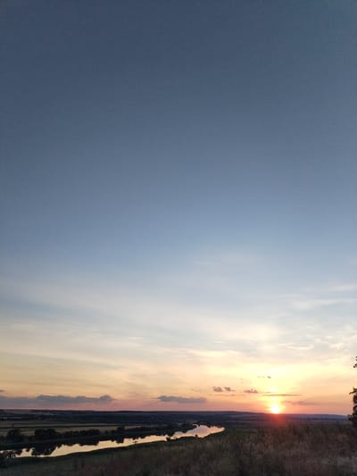 The image captures a serene sunset over a rural landscape in Bilousivka, Ukraine. The sky transitions from a pale blue at the top to soft oranges and pinks near the horizon, with scattered wisps of clouds. The sun is a bright, glowing orb just above the distant treeline, casting a warm reflection on a wide, meandering river that curves through the scene. The riverbanks are lined with dark trees and dense vegetation, contrasting with the lighter, cultivated fields visible in the background. The foreground is dominated by dry, tall grass and foliage, suggesting a natural, untamed environment. There are no people or discernible man-made structures in the immediate view, creating a sense of peaceful isolation and natural beauty.