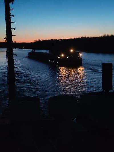 At dusk in Badozhskoi Pogost, Russia, a barge travels along a canal. The sky transitions from blue to orange near the horizon. Dark silhouettes of trees line the canal banks. The barge is illuminated by multiple lights, creating reflections in the water. In the foreground, a dark metallic structure with ladder rungs is positioned to the left, and a post is positioned to the right. Below these is dark indistinct machinery. The water shows the turbulence caused by the barge's passage.