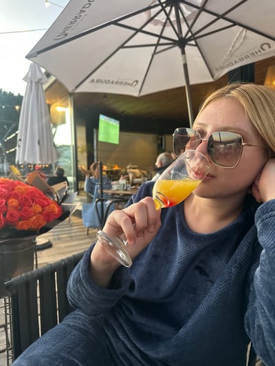 A woman in sunglasses and a blue robe sips a mimosa. A white umbrella labeled "The Boathouse" provides shade in an outdoor setting. Roses in orange and pink hues decorate the table. Other patrons sit at tables in the background. A television screen shows a soccer game in the back.