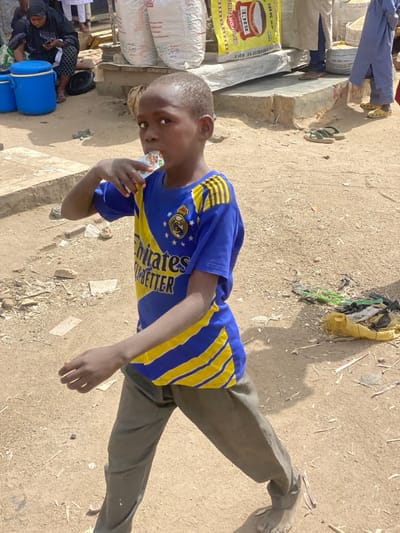 A young Black male child, approximately 5-8 years old, is shown standing in an unpaved outdoor area in Rijiyar Lemo, Nigeria. He is positioned slightly left of center, looking directly at the viewer, with a small, light-colored packaged drink held to his mouth by his right hand. The child wears a blue and yellow striped short-sleeved t-shirt featuring "Emirates FLY BETTER" text and a Real Madrid crest, along with olive green trousers, and is barefoot. The ground is composed of light brown dirt or sand with scattered small debris. In the left background, a woman wearing a dark abaya or hijab is seated on the ground, looking at a smartphone, adjacent to two blue plastic containers. To the right, a raised concrete platform holds several large white sacks, one clearly marked as "FORTUNE RICE." Visible near the platform are the lower legs and feet of multiple adults, some wearing sandals. A pair of green flip-flops rests on the ground. Further right, a yellow sack containing various refuse items is present on the dirt. The scene is brightly illuminated by natural daylight.
