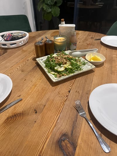 A rustic wooden dining table is set with plates, cutlery, and a salad. The salad, served on a decorative rectangular plate, features arugula, sliced pears, and walnuts. To the right of the salad is a small white bowl filled with sliced butter, and a pair of white salad tongs rests above it. A bottle of "GLAZE" dressing is situated behind the tongs. A lit candle in a green glass holder and a ceramic dispenser are to the left of the salad. In the background, two wooden salt and pepper grinders are visible. A white ceramic basket holding various items, including playing cards and what appears to be sunglasses, sits on the table. The scene is captured from a high angle, looking down onto the table. The location is not Bonville, Australia.