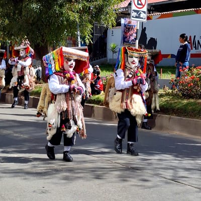 Two individuals in traditional Wanchaq, Peru festival attire, characterized by large, elaborate masks and colorful, layered clothing adorned with fur and tassels, are walking in a procession on a paved street. The masks feature white faces with painted black mustaches and eyes, and are topped with ornate, multi-colored headdresses. Their outfits include white shirts, decorative vests, and dark pants tucked into black boots. They are accompanied by other figures partially visible in the background, and the scene is set against a backdrop of trees, a building with a mural, and a street. Red flowers and green grass are visible along the side of the street, and shadows from trees are cast across the pavement. A girl in a blue sweater and jeans stands observing the procession.