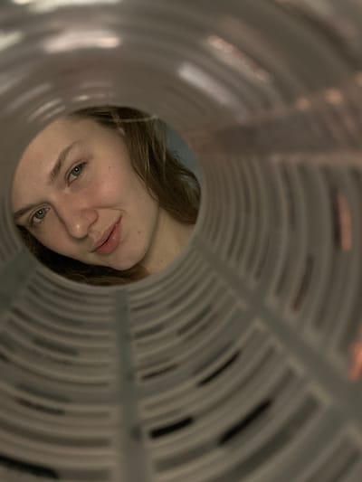 The image captures a female subject looking directly into the lens from an obscured perspective, framed by the interior of a translucent, ribbed cylindrical object. The subject is a light-skinned individual with light brown, wavy hair framing her face. Her features include light-colored eyes, defined eyebrows, a small nose, and full lips parted slightly in a subtle smile. Faint freckles are visible across her nose and upper cheeks. The cylindrical object, appearing light grey, features prominent horizontal ribs with periodic vertical gaps along its interior, creating a tunnel-like aperture. The background behind the subject is out of focus, appearing as an indistinct light blue or greenish-blue surface, indicative of an indoor setting. Some blurred light sources or reflections are visible through the upper right portion of the cylindrical object's material. The scene is situated in Tashevka, Russia.