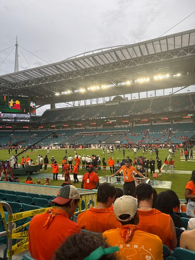 The image depicts a wide view of Hard Rock Stadium located in Miami Gardens, Florida, United States, under an overcast sky with stadium floodlights illuminated. The scene is observed from a lower-tier spectator seat, with yellow "CAUTION" tape visible on a railing in the immediate foreground. Several individuals, primarily seen from behind, occupy teal stadium seats in the foreground, most wearing orange shirts; one individual wears a multi-colored baseball cap and an orange bandana, while another wears a white baseball cap.
On the green artificial turf field below, numerous individuals are gathered. A significant portion wear orange shirts or vests, while others are dressed in white or black attire, including some appearing to be football players in uniforms. A male in an orange polo shirt and black cap stands leaning on a railing at field level, facing the activity. A large camera rig is positioned on the left side of the field.
The multi-tiered stadium seating, primarily teal, extends into the background, with scattered spectators visible. A large video scoreboard on the left displays an animated character labeled "BIG CHEESE," the "UNIVERSITY OF MIAMI HEALTH SYSTEM" logo, and the time "6:41 PM." The stadium's expansive metal roof structure and multiple bright floodlights are prominent against the grey, cloudy sky. A tall, pointed mast structure is visible extending above the roofline.