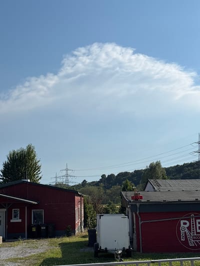 A wide shot shows a residential area in Bochum, Germany. The image features a clear blue sky with a large cloud formation. In the foreground, there are two red buildings, one on the left with a white door and window, and another on the right with a large red banner displaying the text "RIB BOCHUM" within a circular logo. A white trailer is parked between the two buildings. In the background, there are power lines supported by tall metal pylons and a densely wooded hillside. The ground is covered with gravel and grass.
