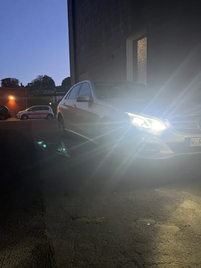 The image depicts a nighttime scene in Wuppertal, Germany. A white Mercedes-Benz, facing the viewer with its headlights on, dominates the foreground. The car is parked under a building with a recessed, illuminated ceiling feature. To the left, the sky transitions from a blue gradient to darkness. Behind the Mercedes, a smaller white vehicle with red accents is visible, possibly an ambulance or service vehicle. The ground appears to be a paved surface, possibly a road or parking area. The scene is brightly lit by the car's headlights and nearby streetlights. A tree line and building structures are vaguely discernible in the background. A lamp post and what appears to be a scooter or small motorcycle are also visible at the bottom of the picture.