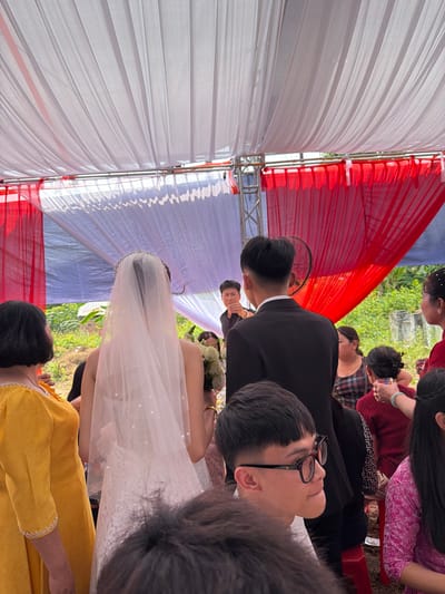 This image, taken in Tay Son Commune, Vietnam, captures a wedding ceremony or reception held outdoors or semi-outdoors under a canopy. The canopy is adorned with large drapes of white and red fabric, creating a festive backdrop.
In the midground, the bride is prominent, viewed primarily from behind. She wears a traditional white wedding gown with a sheer veil extending down her back and a tiara on her head. She holds a bouquet of white flowers. The groom stands nearby, dressed in a black suit and white shirt, with his back mostly to the camera.
Several guests are visible around the couple. In the corrected foreground, a young man with short dark hair and glasses looks up and to the side. Another guest in a bright yellow dress is partially seen in the foreground. Other guests, some in traditional Vietnamese attire or formal wear, are scattered throughout the scene. One guest is seen looking down at a mobile phone. A small electric fan is visible near the groom, suggesting warm conditions.
The background features lush green foliage, indicating an outdoor setting. The bright, clear lighting suggests the event is taking place during the daytime. The overall mood appears celebratory, consistent with a wedding. There is no visible text in the image.