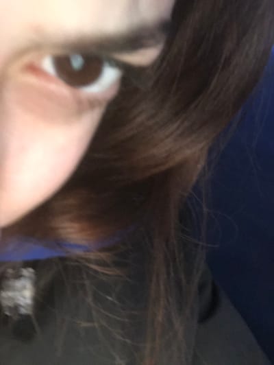 A close-up shot focuses on a woman's face, specifically her eye and surrounding skin. Her brown eye is prominent, with a visible white reflection. Dark brown hair frames the face, some strands appearing lighter. The background is blurred, with hints of dark blue and black clothing. The image suggests a candid or personal moment, captured with a shallow depth of field.