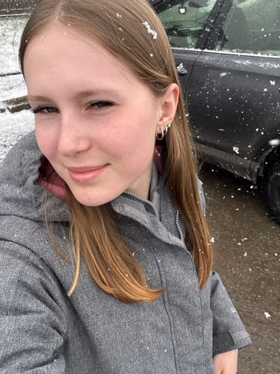 The photograph features a young woman with long, light brown hair partially covered in snowflakes. She is wearing a gray jacket and has multiple silver hoop earrings in her left ear. Snowflakes are visible in the air and on her hair and jacket, suggesting it is snowing. A black car is partially visible in the background. The location is not definitively identifiable as Lunezhki, Russia without additional visual or contextual clues.