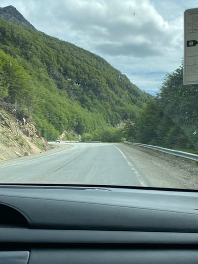 A view from inside a vehicle shows a winding road next to a steep, green mountainside. The road is bordered by a guardrail on one side. Trees and vegetation cover the mountain, and the sky is partly cloudy. The car's interior, including the door panel, is visible on the right side of the image. A label is attached to the windshield on the upper left.