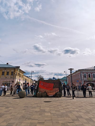 The image shows a staged historical exhibit in the city of Uglich, Russia. The exhibit appears to be a small red brick structure decorated with camouflage netting and sandbags. A red banner with a hammer and sickle symbol hangs on the structure. Text on the banner reads "150th Order of Kutuzov ISTR. IPRIZ DIV 70 S.V. Z U.A. 16.02". A metal fence is positioned in front of the exhibit, separating it from the pedestrian area. A group of people, including men and women of varying ages, are standing behind the fence, some leaning on it as they observe the display. The background features a paved square surrounded by multi-story buildings with architectural details. The sky is partly cloudy, with visible contrails from aircraft. Street lamps are placed along the square. Two commercial signs visible in the background, one reading "АлинУМ" and "ВРЕМЯ ПО УГЛИЧ" suggesting this location is a central square.