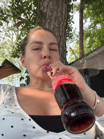 This is a portrait of a young woman drinking from a glass Coca-Cola bottle. She is outdoors, with a tree trunk and lush green foliage visible in the background, suggesting a natural, possibly wooded setting. The lighting indicates it is daytime, with dappled sunlight filtering through the leaves.
The woman is holding the bottle to her lips, tilting her head back slightly as she drinks. Her facial expression is relaxed, with her eyes partially closed and her lips pursed around the bottle opening. She has dark nail polish on her fingers, and a silver bracelet with a heart charm adorns her wrist. She is wearing a white top with black polka dots.
Visible text on the Coca-Cola bottle label is in what appears to be Bosnian or a similar South Slavic language. The text mentions "Osvježavajuće bezalkoholno piće" (Refreshing non-alcoholic drink) and provides contact information for Coca-Cola in Hadžići, Bosnia and Herzegovina. The location context provided suggests the photo was taken in Ćusine, Bosnia and Herzegovina. The overall impression is a casual, refreshing moment enjoyed outdoors on a pleasant day.