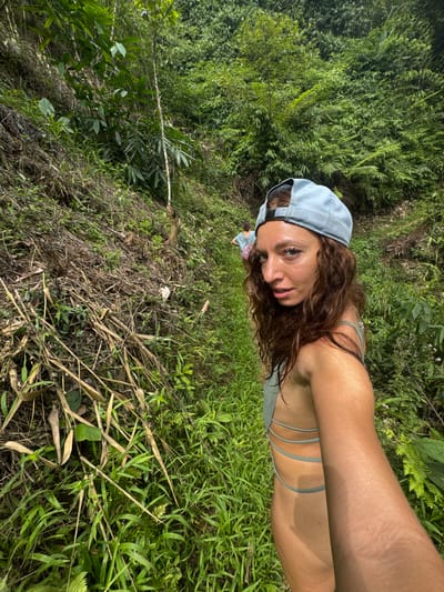 A young adult female takes a selfie outdoors in Suối Giàng, Vietnam. She wears a light blue baseball cap and a bathing suit top. The background consists of dense vegetation and tall grass.