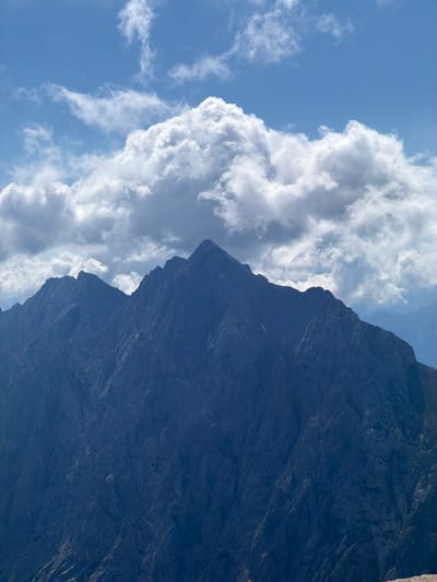 This is a medium shot, taken from a low angle, of a dramatic mountain landscape under a blue sky with cumulus clouds. The image is oriented vertically, with the mountain dominating the right side of the frame and the sky with clouds filling the left. The mountain appears to be a sheer, dark, rocky face, with deep shadows and visible texture indicating its rugged surface. There are no people or man-made objects visible.
The scene suggests a clear day with fair weather, as indicated by the bright blue sky and fluffy white clouds. The lighting suggests it is daytime, likely mid-morning or afternoon, given the brightness and the shadows on the mountain. The location is identified as Wildermieming, Austria, which is known for its mountainous terrain, consistent with the visual information in the image. The overall mood is one of natural grandeur and rugged beauty. There is no visible text in the image.