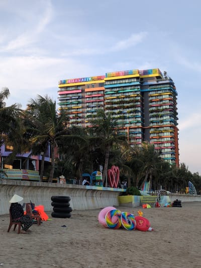 In the coastal city of Hoằng Tiến Commune, Vietnam, a vibrant, multi-colored hotel named "Ibiza Party Resort" dominates the skyline, its facade adorned with a spectrum of pink, yellow, blue, and green hues. The resort features numerous balconies, many of which are lined with lush greenery, suggesting a tropical setting. Surrounding the resort are tall palm trees, their fronds creating a silhouette against the pale blue sky dotted with wispy clouds.

In the foreground, the sandy beach is scattered with various objects. A woman wearing a conical hat and traditional patterned clothing sits on a chair, appearing to be engrossed in her phone. Beside her are two empty chairs and a bright orange life vest. A stack of four black inner tubes rests on the sand. Further along the beach, several inflatable toys are strewn about, including colorful ring floats with rainbow patterns and a red, rocket-shaped inflatable. In the middle ground, a low concrete wall separates the beach from a promenade area, where more palm trees grow. A few people are visible on the promenade, including a couple sitting on steps and a person in the distance near some playground equipment. The overall scene captures a relaxed atmosphere on a beach adjacent to a lively, architecturally distinct resort.