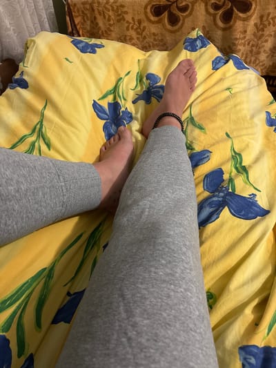 The image shows a close-up view of a person's legs and feet resting on a yellow bedsheet with a blue floral pattern. The person is wearing grey sweatpants and has a black beaded anklet on their left ankle. The background includes parts of a curtain and a patterned fabric, suggesting an indoor domestic setting. There is no indication of the location being in the city of Zgorigrad, Bulgaria.
