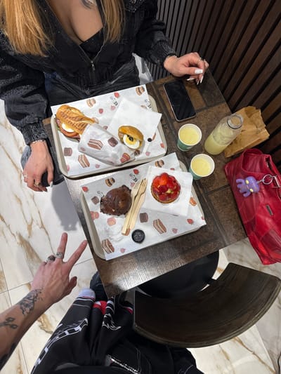 This image captures an overhead view of a café table in Asnières-sur-Seine, France, occupied by two individuals. One person, positioned at the top of the frame, is partially visible from the chest up, possessing long blonde hair and wearing a black sequined jacket over a black top. Multiple rings adorn her fingers, and her left hand rests near a black smartphone lying face-up on the dark wooden table. The second individual's left arm and upper thigh are visible from the lower-left corner; this person wears dark patterned clothing, displays tattoos on the forearm, rings on the fingers, and makes a peace sign with two fingers.
Two rectangular serving trays, each lined with white paper featuring pastry and strawberry motifs, are on the table. The upper tray holds a grilled sandwich (panini) wrapped in paper and a smaller bun containing a sliced hard-boiled egg and an olive. The lower tray presents a dark round pastry (possibly a chocolate muffin), a strawberry tart, a small lidded black cup containing a dark substance, and a wooden fork and knife set.
Beverages include two light yellow liquids in green-rimmed paper cups and a glass bottle filled with the same liquid. A red handbag with a purple octopus keychain rests on the table to the right of the beverages. The immediate environment includes a dark vertically slatted wooden wall behind the table and a light-colored marble-patterned floor. A dark chair back is visible in the lower right.