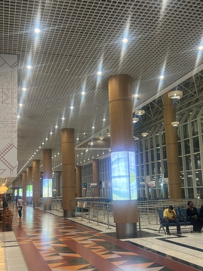 This media file depicts a brightly lit, modern interior space, strongly suggesting an airport terminal or a large public waiting area in Addis Ababa, Ethiopia. The scene is characterized by contemporary architecture and a sense of spaciousness.

The ceiling features a distinctive white grid pattern, from which numerous small, recessed lights emit bright beams, creating visible lens flares across the upper half of the image. Several large, horizontal, cylindrical structures, wrapped in light-colored wooden slats, extend through the upper portion of the hall. Integrated into these wooden cylinders are decorative, woven light fixtures resembling large lanterns.

One of these prominent cylindrical structures incorporates a large rectangular digital display screen, which shows a landscape image, possibly of mountains or natural scenery. Clearly visible text on this screen reads "Bale Mts, Oromia, Ethiopia" and "Photo: [unclear]". Further down the hall, similar cylindrical structures and screens can be seen.

The flooring is a combination of light-colored, possibly polished, tiles and a section with a striking geometric pattern in shades of brown, orange, and white. Along the right side of the image, multiple individuals are seated on public benches, appearing to be waiting. One person in a yellow shirt and dark pants, and another in a dark top, are prominent in the mid-ground. Other people are scattered throughout the extensive hall.

Metal barriers and structural elements are also visible, delineating various areas. Signs attached to pillars or walls clearly indicate "ZONE 1", "ZONE 3", "ZONE 5", and "C2", typical of a large public facility. The lighting is entirely artificial, contributing to a clean and bright indoor atmosphere. The activity observed is primarily people waiting in a public setting.