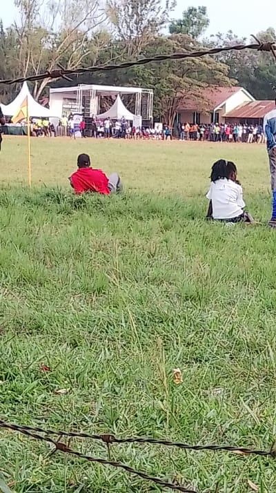 The image depicts an outdoor public gathering in Siaya Township ward, Kenya, viewed from a slightly elevated position behind a barbed wire fence. The foreground features a two-strand barbed wire fence running horizontally across the bottom, in front of a wide expanse of green grass. In the left-center foreground, an individual in a red shirt and grey pants is lying prone on the grass, facing towards the left. To their right, another person with dark hair, wearing a white shirt and dark trousers, is seated on the grass, facing away from the camera. A yellow flag on a pole is visible in the mid-left, partially obscured.
In the mid-ground, a large group of people is assembled around a white outdoor stage structure with prominent speaker systems on either side. Multiple white canopy tents are erected around the stage, one on the left displaying an orange banner. The crowd is composed of individuals standing and seated, many in light-colored attire, facing the stage. The background includes several light-colored buildings with reddish-brown corrugated roofs, situated amidst numerous mature trees with green foliage, under a bright sky.