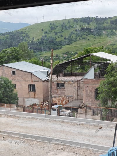 This media file captures a rural scene in Bazum, Armenia, likely during the daytime under overcast or hazy weather conditions, as suggested by the diffused light and cloudy sky. The foreground features a newly constructed concrete pathway or road with gravel and debris, indicating ongoing development or maintenance.

Dominating the mid-ground are two unfinished or dilapidated buildings constructed from stone and concrete, with corrugated metal roofs. The building on the left is a two-story structure with a peach-colored facade and a light gray metal roof. It has visible windows, some boarded up, and a large, old-fashioned garage door. The building on the right appears to be a more open structure, possibly a workshop or shed, with a skeletal framework and a white metal roof. Both buildings show signs of age and disrepair, with rust evident on some metal elements and weathered walls. A wooden utility pole stands prominently between the two structures. Behind the buildings, a chain-link fence with rusted elements separates the property from the road. Beyond the fence, there are glimpses of old vehicles, including what appears to be a light-colored van or car partially obscured.

The background is characterized by a lush, green, rolling hillside dotted with trees and shrubs. Two tall electrical transmission towers are visible on the crest of the hill, indicating the presence of power infrastructure. The overall impression is one of a developing rural area, where modern construction is juxtaposed with older, weathered structures and the natural landscape. There is no visible text in the image.