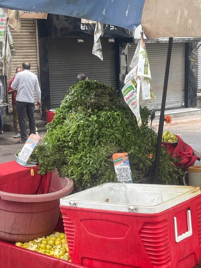 In Ahmedabad, India, a street vendor is preparing a beverage. A large pile of fresh mint leaves is central to the scene, with a red cooler in the foreground, filled with ice and lemon slices. In the background, two men are standing near a closed storefront. A sign on the storefront displays a phone number, likely for business contact. A blue and tan umbrella provides shade over the stall.