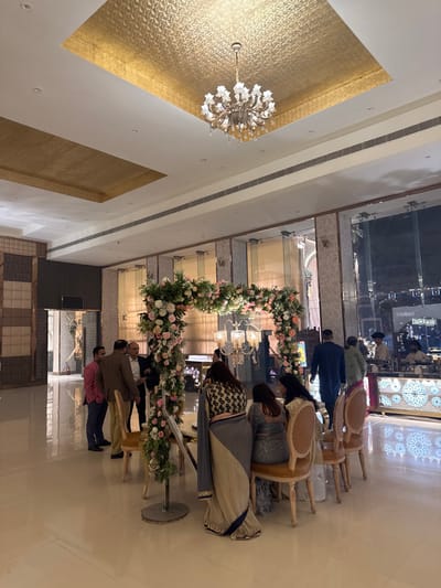 A group of people stands before a floral archway indoors. Chairs line the space behind them. Above, a golden panel with a chandelier hangs from the ceiling. Large glass windows reflect interior scenes. The scene suggests a formal event or gathering in a spacious hall.