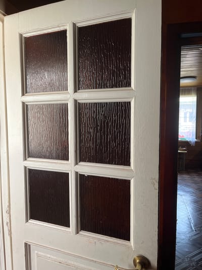 The image shows an interior view of a white wooden door with six rectangular panes of textured, dark brown glass. The door is slightly ajar, revealing a hallway or room beyond. The door frame is also white and appears to be painted. The glass panes have a ribbed pattern, creating a distorted view of the space behind the door. In the background, a wooden door frame is visible, leading to a room with wooden walls and a window covered by a white lace curtain. The floor in the background appears to be dark and patterned. A brass door handle is visible on the lower section of the door. The image is taken from a slightly low angle. This scene is located in Kozitsino, Russia.