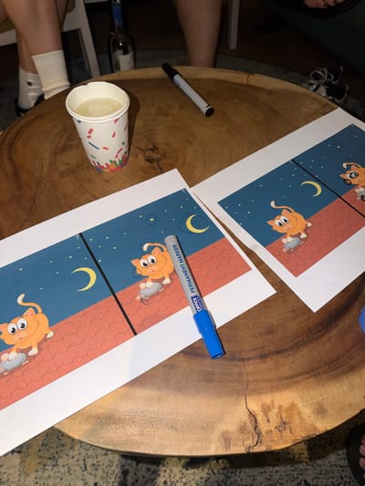 The image depicts a close-up of a wooden table with several items on it. In the foreground, there are two sheets of printed paper featuring a cartoon of an orange cat on a rooftop under a starry night sky with a crescent moon. A blue permanent marker with the label "OFFICE PERMANENT MARKER" is positioned across the right sheet of paper. To the left of the papers, there is a white paper cup with a colorful confetti pattern, filled with a light-colored liquid. Further back, a partially visible bottle with a dark neck and a white label, possibly a wine bottle, stands on the table. The legs of two individuals are visible on the left side of the image; one is wearing white socks and the other is wearing dark shoes. The overall environment appears to be indoors, with the wooden table being the central focus. There is no indication of the location being in Heijen, Netherlands.