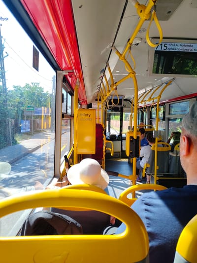 The image shows the interior of a bus in Jaworowa, Poland, looking forward. The interior is characterized by bright yellow handrails and poles against a predominantly white ceiling and red trim along the windows. Outside the window, a street scene with trees and buildings is visible, suggesting a daytime setting. Several passengers are seated on the bus, including a person in the foreground facing away from the camera, wearing a blue shirt and a grey cap. Other passengers are visible further inside the bus, some looking forward or towards the windows. A yellow ticket machine is mounted on the wall to the right, labeled "bilety tickets". Digital displays showing route information are also visible overhead, with one displaying "715 -> P+R Al. Krakowska". The overall impression is of a typical public transportation environment.
