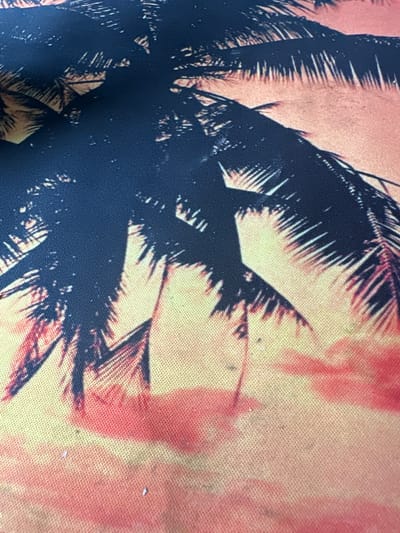 This image is a close-up shot of a fabric with a printed design. The design features silhouettes of palm fronds against a gradient background that transitions from a light yellow to a peachy-orange, with some areas of darker pinkish-red. The palm fronds are a deep navy blue. The fabric has a visible woven texture. There are no people or specific locations in the city of Sinegorye, Russia depicted in this image.