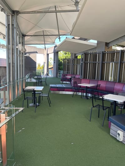This image depicts an outdoor or semi-outdoor dining/lounge area, likely part of a commercial establishment in Kelvin Grove, Australia. The area features a vibrant green artificial turf floor.
The primary subjects are the numerous pieces of outdoor furniture. Along the left side of the frame, a long stretch of cushioned bench seating is visible, alternating between sections upholstered in a rich purple/maroon and others in a light grey. In front of these benches are multiple small, square, white-topped tables paired with black metal chairs. Many of these chairs are stacked neatly, suggesting the area might be closed, preparing to open, or being cleaned.
Overhead, a large white tensile fabric canopy, supported by metal beams and cables, provides shade to the left portion of the space. Several lights and fixtures are attached to this canopy structure. A glass railing or partition runs along the right side of the viewing area, reflecting some of the surroundings. Beyond the benches, there's a slatted metal wall or barrier, and through its openings, green trees and distant building structures are visible under bright, clear daylight.
No people are present in the scene, implying a quiet or off-peak moment. The bright lighting indicates it is daytime, likely sunny. A white rectangular object with two circular openings, possibly an electrical box or vent, is situated on the artificial turf on the far right. A faint number "7" can be seen on a panel near the slatted wall. The scene is orderly, clean, and ready for guests.