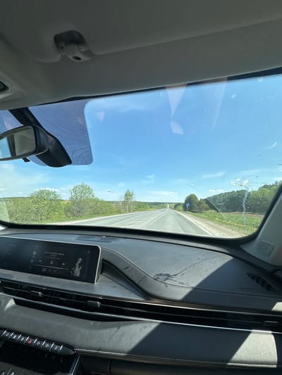 The image depicts a view from inside a car, looking out onto a straight road with green vegetation on either side. The dashboard of the car is visible, featuring a digital display showing a song title in Russian, "A мы не ангелы, парень" by OSTRA. Various buttons and controls are also present on the dashboard. The windshield has some smudges and marks, partially obscuring the view of the blue sky and distant trees. A rearview mirror is seen in the lower left corner of the frame. The location is likely Postol, Russia given the song title in Russian.