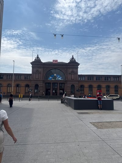 The image shows the Bonn Hauptbahnhof train station in Bonn, Germany. The building is made of brown brick and has a large arched entrance with a clock above it. The DB logo is visible on the building. There are several people in front of the station, some of whom are sitting or lying down on a large black structure. The sky is blue with scattered clouds, suggesting it is daytime and the weather is pleasant. The ground is paved with grey tiles.