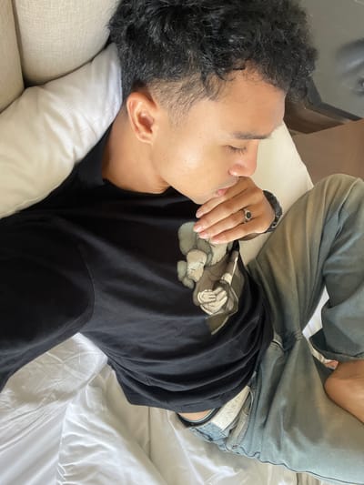 A male individual is depicted lying on a bed in the city of Kayuambon, Indonesia. He has dark, curly hair with a faded cut on the sides, and his eyes are closed or cast downwards. He is wearing a black long-sleeved t-shirt featuring a graphic design of a stylized character with crosses for eyes, and light-wash denim jeans. A silver-colored ring with a dark oval stone is present on the ring finger of his left hand, which rests near his chin. A dark, textured wristband is visible on his right wrist. He is positioned against a white pillow, which abuts a light-colored, upholstered surface, likely a headboard or sofa back. White bedsheets are visible beneath him, and a darker surface is partially observable in the background.