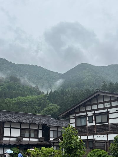 This detailed image captures a panoramic view in the city of Shirakawa, Japan, under an overcast sky. Dominating the upper two-thirds of the frame are lush, densely forested mountains, appearing vibrant green with shades of dark green, especially at the lower elevations. Wisps of white mist and fog cling to the mid-slopes of the mountains, partially obscuring the trees and creating a serene, almost mystical atmosphere. The sky is a uniform, light grey, heavily laden with clouds, suggesting a recent or ongoing rainfall.

In the foreground, partially cropped on the right and extending across the bottom left, are two traditional Japanese-style buildings with distinctive architectural features. The building on the right features dark wooden beams forming a grid pattern against white plaster panels, characteristic of old Japanese architecture. It has dark tiled roofs and multiple windows, some with closed curtains. The building on the left shares a similar aesthetic with dark wood framing and white walls, featuring several windows and a dark, intricately patterned roof. Green foliage and small trees are visible around and between the buildings, adding to the natural setting. Below the eaves of the leftmost building, a few individuals are partially visible, one holding a light blue umbrella and another a pink umbrella, indicating rainy weather. The overall scene conveys a tranquil, traditional Japanese rural landscape amidst a misty mountain backdrop.
