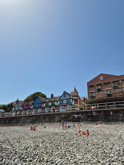 The image captures a scene at Penarth Beach in the United Kingdom. A pebbly beach is adjacent to a concrete sea wall. People are lying on the beach, sunbathing. Behind the sea wall are buildings, varying in height and style. One building is made of red brick with a checkerboard emblem. There are colorful buildings behind that with timber frames and gabled roofs. The sky above is blue.