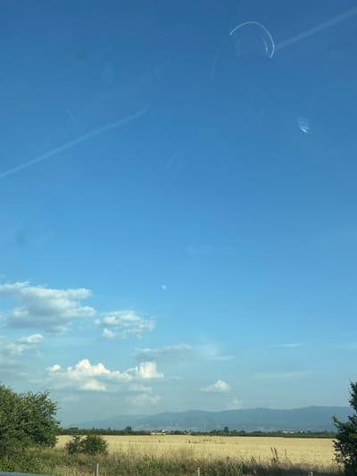 The image is an outdoor shot of a rural landscape under a bright blue sky with scattered white clouds. The orientation of the image is vertical. The foreground on the left is dominated by the clear blue sky, showing some reflections and smudges, possibly from a window or lens. In the sky, a small, faint white circle is visible, which could be the moon.
The right side of the image shows a field of dry, yellowish grass, likely a harvested crop, stretching towards a distant line of hazy mountains. A few trees and bushes are visible along the edge of the field and in the foreground. A fence post is also visible on the far right. In the distance, some buildings can be seen nestled at the foot of the mountains. The sunlight suggests it is daytime, likely late morning or afternoon, and the weather is clear and sunny. The scene depicts a tranquil rural setting. There is no visible text in the image.