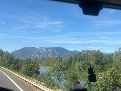 The image captures a scenic view from a vehicle, likely driving along a road adjacent to a river. In the foreground, a highway with a white line marking the lane is visible on the left. A metal guardrail runs along the edge of the road, separating it from the natural landscape. The middle ground is dominated by a wide, calm river with a turquoise hue, reflecting the bright sky. Dense green trees line the riverbanks, creating a lush forest. In the background, a majestic mountain range rises, its rocky slopes and peaks casting shadows under a clear blue sky dotted with wispy white clouds. The sunlight suggests it is daytime, likely midday or early afternoon. The overall atmosphere is serene and picturesque, evoking a sense of travel and natural beauty. There are no people visible, but a small black electronic device is hanging from the windshield inside the vehicle. No text is present in the image. The location context provided is Barraux, France.