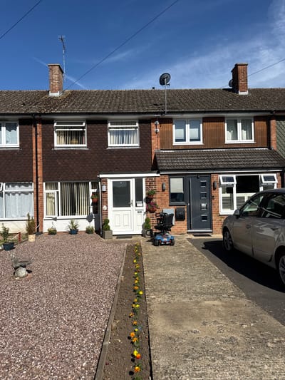 The image depicts a row of semi-detached houses in West Oxfordshire, United Kingdom. The houses are two stories tall, with brick and brown tiled exteriors. White-framed windows are visible on each house. A grey car is parked on the driveway of the house on the right. A blue mobility scooter is parked in front of the adjacent house, addressed as "45". The house on the left is addressed as "43". Flowerbeds line the driveways, adding color to the scene. The sky above is blue with scattered clouds. Chimneys and satellite dishes are affixed to the roofs. The foreground shows a gravel area and flowerbed.