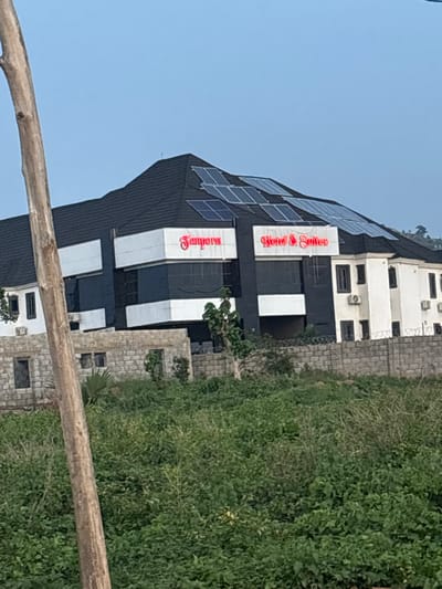 The image shows a modern building with a dark facade, white accents, and solar panels on its angled roof. The building appears to be a hotel or guesthouse, with "Hotel & Suites" and "Sempore" written in red lettering on the facade. The building is surrounded by lush green vegetation and a stone wall. The sky is clear and blue, suggesting it is daytime. The image is taken from a slightly elevated angle, looking across a wooden beam in the foreground. The overall scene depicts a well-maintained establishment in a tropical or sub-tropical environment. No people are visible in the image.