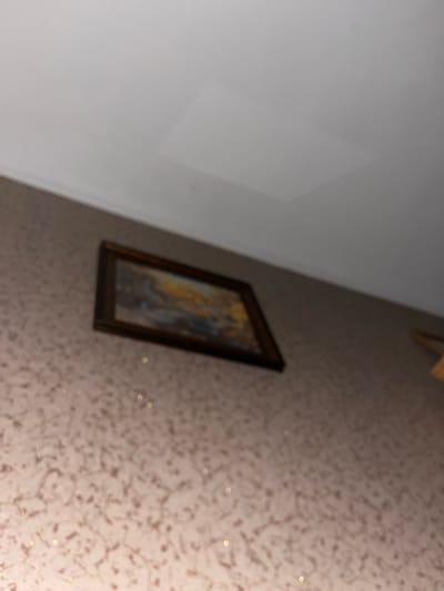 The image shows a landscape painting in a wooden frame hanging on a wall with patterned wallpaper. The ceiling above is white and appears to have some discoloration or a lighter patch. The image is out of focus, making it difficult to discern specific details of the painting or the wallpaper pattern. There is no discernible information about the location in the city of Ponebel, Ukraine, or any people, actions, or interactions.