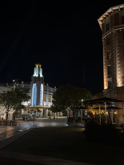 A night scene features two contrasting architectural styles. At the top, a brick building with ornate details is partially visible against the dark sky. Below, a modern building with illuminated features, including a blue-lit vertical element, stands prominently. Trees frame the modern structure, and a paved area with seating and sparse lighting suggests a public space. The composition is shot from a low angle, accentuating the height and scale of the buildings.