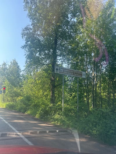 The image shows a road in Posyolok Imeni Morozova, Russia. A signpost with a directional arrow points to the right, indicating "КРЕПОСТЬ ОРЕШЕК" and "ORESHEK fortress" at a distance of 2.0 kilometers. The road is paved and has a white line marking the lane. On the left side of the road, there is a traffic light showing red. Dense green foliage and trees line the right side of the road. The sky is clear and blue.